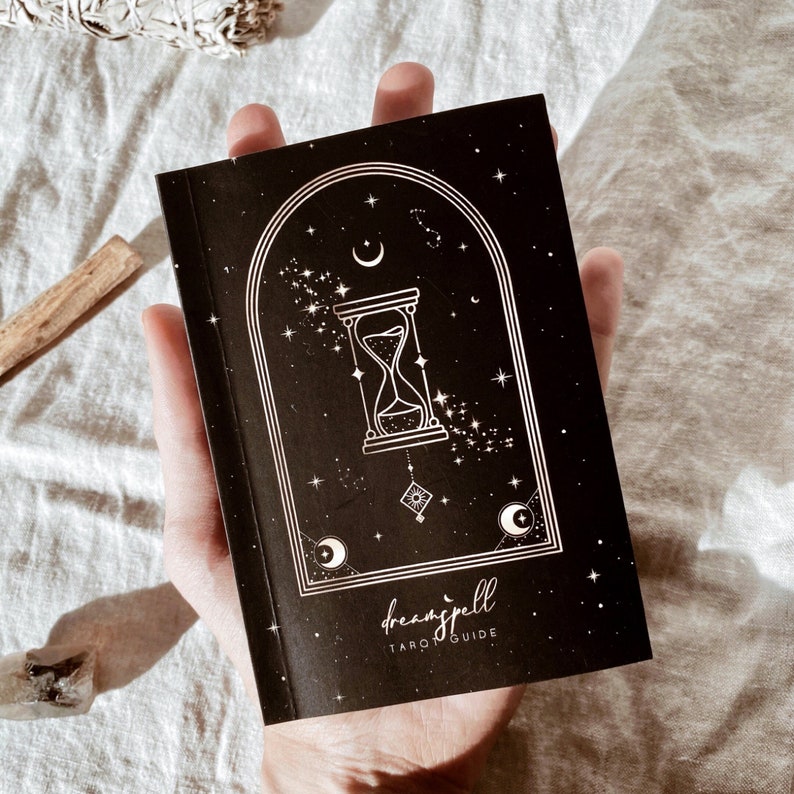 Puede incluir: Un libro de gu&iacute;a de tarot negro con una ilustraci&oacute;n blanca de un reloj de arena, estrellas y una luna creciente. El t&iacute;tulo "dreamspell TAROT GUIDE" est&aacute; impreso en blanco en la portada.