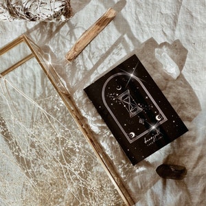 Puede incluir: Una gu&iacute;a de tarot negra con detalles en dorado. La portada presenta una ilustraci&oacute;n de un reloj de arena, estrellas y una luna creciente. El texto en la portada dice "dreamspell tarot guide".