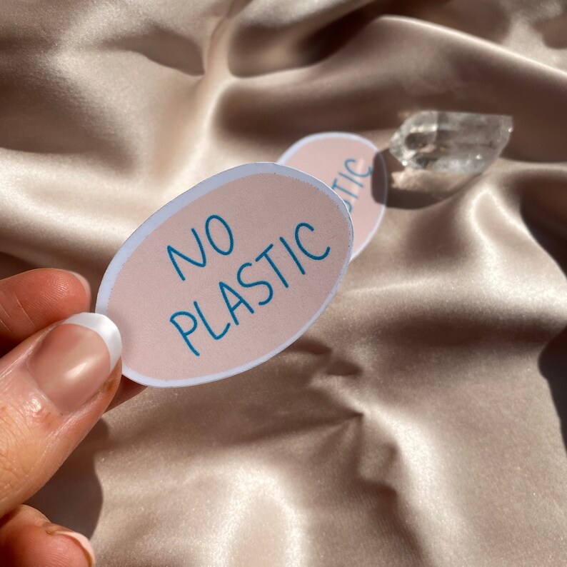 May include: Two oval stickers with the words "NO PLASTIC" in teal lettering. The stickers have a light pink background and a white border. The stickers are on a satin fabric.
