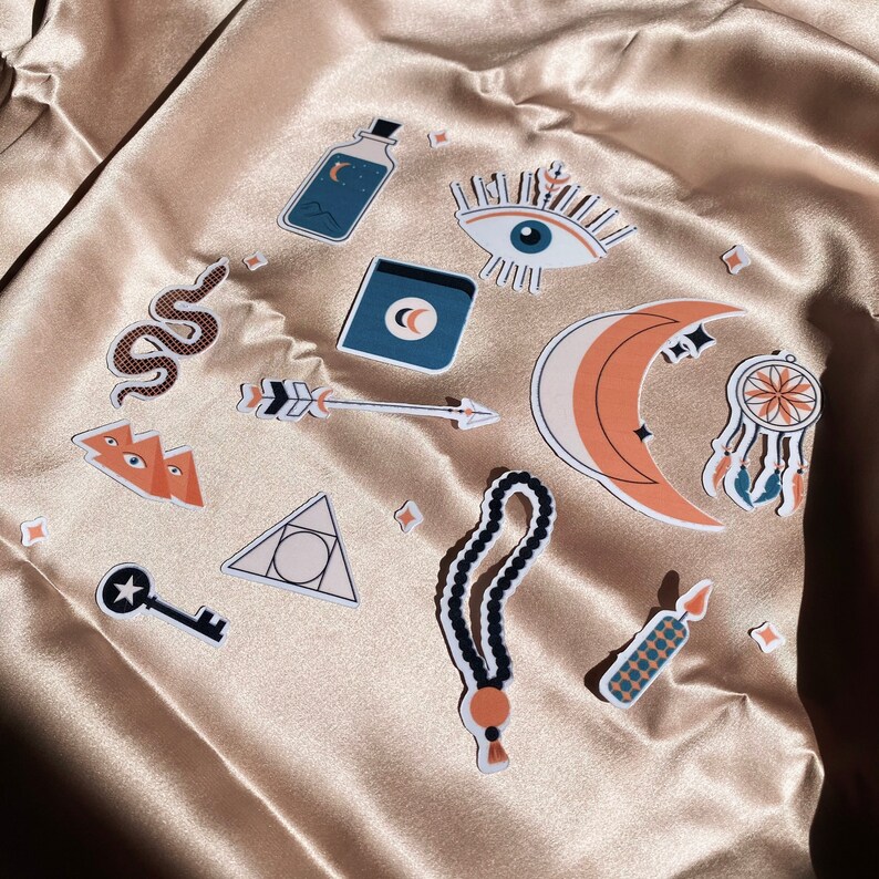 May include: A set of 11 stickers featuring various mystical and spiritual symbols, including a crescent moon, an eye, a snake, a key, a dreamcatcher, an arrow, a triangle, a bottle, and a candle. The stickers are on a beige fabric background.