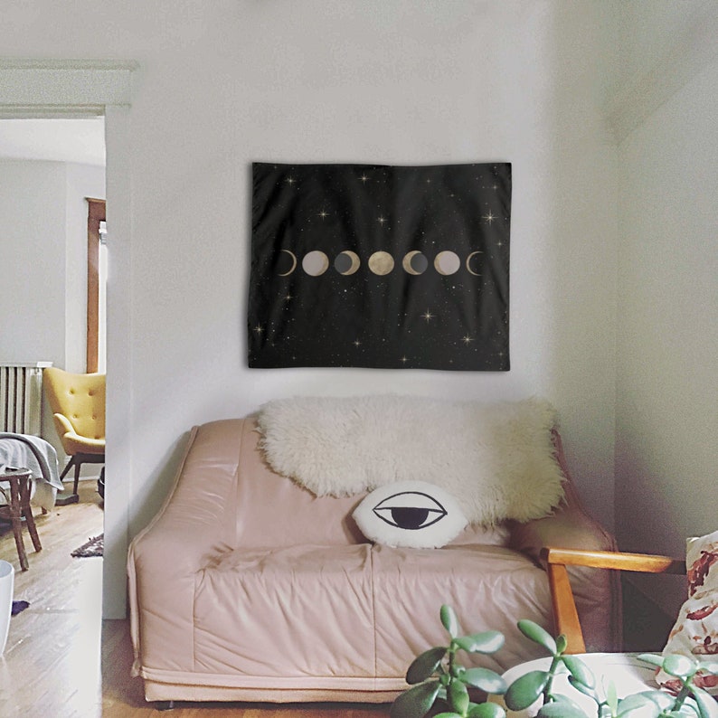 May include: A black tapestry with a gold and white moon phases design on a black background with white stars. The tapestry is hanging on a white wall above a pink couch with a white fur throw and a black and white eye pillow.