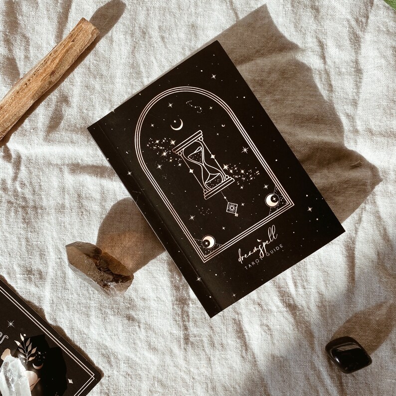 Puede incluir: Un libro negro con una ilustraci&oacute;n dorada de un reloj de arena, estrellas y una luna creciente. El t&iacute;tulo del libro es "dreamspell tarot guide".