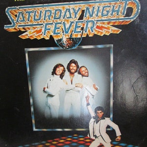 May include: A vintage vinyl record album cover for the original movie soundtrack of "Saturday Night Fever". The cover features a disco ball and a group of four men in white suits, with one man in a white suit dancing on a disco dance floor.