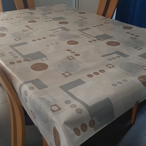 Pode incluir: Uma toalha de mesa branca com um padrão geométrico de círculos e quadrados marrons e cinzentos. A toalha de mesa está sobre uma mesa de madeira com duas cadeiras.