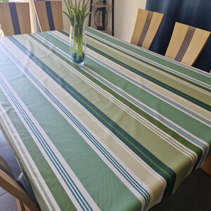 Op de afbeelding: Een rechthoekig tafelkleed met een verticaal streeppatroon in verschillende tinten groen, waaronder salie, olijf en donker bosgroen, geaccentueerd met dunne witte en donkerblauwe lijnen, dat een eettafel met houten stoelen bedekt.