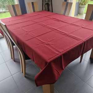 Peut inclure: Une nappe rectangulaire rouge foncé recouvre une table à manger en bois. La table est entourée de six chaises en bois avec des cadres clairs et des accents foncés. La scène se déroule dans une pièce bien éclairée avec vue sur la verdure.
