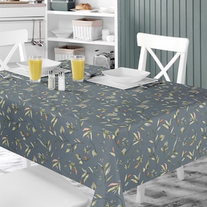 Peut inclure: Une nappe bleue avec un motif floral composé de petites feuilles et fleurs délicates dans des tons de vert, jaune et orange. La nappe est posée sur une table avec deux chaises blanches et une étagère blanche en arrière-plan.