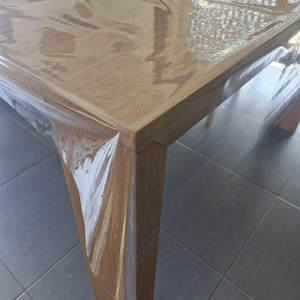 Könnte beinhalten: Ein rechteckiger Holztisch mit hellbrauner Oberfläche, in transparente Folie verpackt. Der Tisch hat ein schlichtes, modernes Design mit quadratischen Beinen und einer glatten Oberfläche. Der Tisch ist wahrscheinlich neu und unbenutzt.