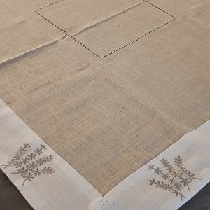 Può includere: Un tovaglia di lino bianco con un centro beige e disegni floreali ricamati intorno al bordo. La tovaglia ha un quadrato cucito al centro.
