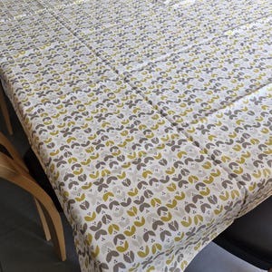 Peut inclure: Une nappe blanche avec un motif répétitif de feuilles brunes et jaunes. La nappe est posée sur une table avec deux chaises en bois.