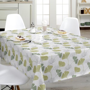 Op de afbeelding: Een rechthoekig tafelkleed met een herhalend patroon van ginkgo biloba-bladeren in tinten saliegroen, olijf en lichtgrijs op een witte achtergrond. De stof ligt over een eettafel die gedekt is met witte borden, kopjes en bestek.