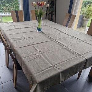 Peut inclure: Une nappe rectangulaire beige recouvre une table à manger en bois. Un vase en verre transparent avec des tulipes colorées est au centre. Des chaises en bois avec une bande sombre au dos entourent la table. La pièce a de grandes fenêtres.