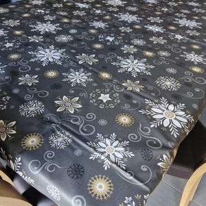 Peut inclure: Une nappe grise avec un motif de flocons de neige blancs et dorés. La nappe est sur une table avec deux chaises en bois.