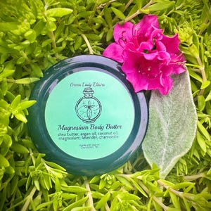 May include: A green jar of magnesium body butter with a white label that reads "Green Lady Elixirs" and "Magnesium Body Butter" with a list of ingredients: shea butter, argan oil, coconut oil, magnesium, lavender, chamomile. The jar is sitting on a bed of green plants.