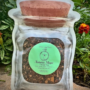 May include: A clear glass jar filled with loose leaf herbal tea, labeled "Autumn Magic Herbal Tea" with a list of ingredients including tulsi, cacao, clove cinnamon, sarsaparilla, and elderberry. The jar is in a white resealable bag with a cork lid.