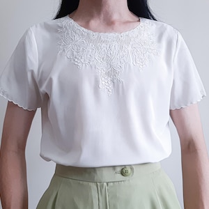 Puede incluir: Una blusa blanca de manga corta con cuello redondo que presenta un intrincado bordado floral blanco y detalles calados. Las mangas tienen un borde festoneado. La blusa se combina con pantalones de color verde claro con un botón visible.