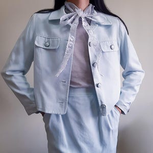 K&ouml;nnte beinhalten: Hellblaues Jacken- und Rock-Set. Die Jacke hat zwei Vordertaschen und Knopfverschl&uuml;sse. Ein wei&szlig;er Spitzenschal wird mit dem Outfit getragen. Der Rock ist in passender hellblauer Farbe gehalten.