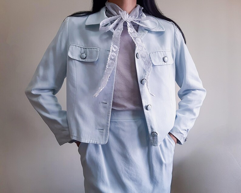 K&ouml;nnte beinhalten: Hellblaues Jacken- und Rock-Set. Die Jacke hat zwei Vordertaschen und Knopfverschl&uuml;sse. Ein wei&szlig;er Spitzenschal wird mit dem Outfit getragen. Der Rock ist in passender hellblauer Farbe gehalten.