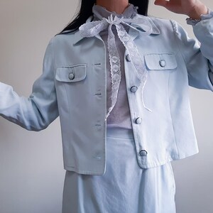 K&ouml;nnte beinhalten: Ein hellblauer zweiteiliger Anzug. Die Jacke hat zwei Vordertaschen und Knopfverschl&uuml;sse. Das Outfit umfasst einen hellblauen Rock und eine transparente, hellfarbene Bluse mit einer Spitzenschleife.