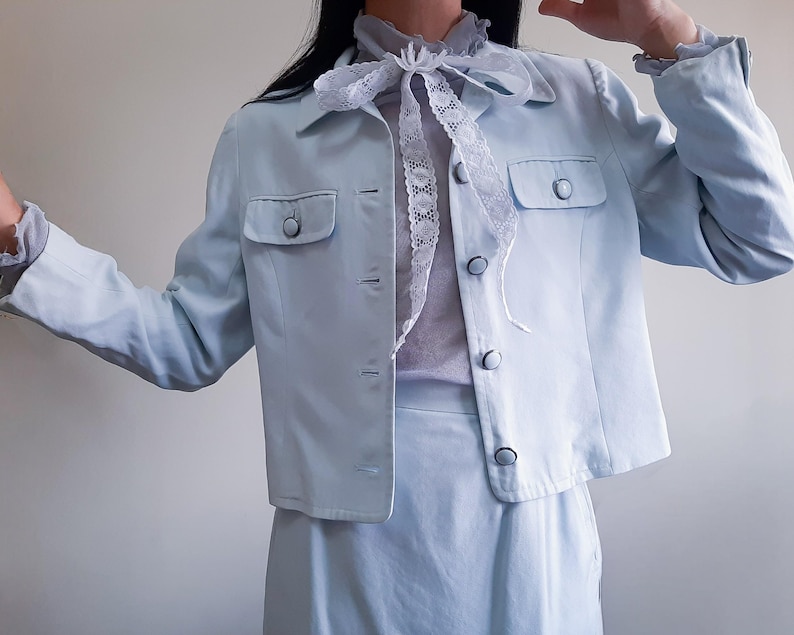 K&ouml;nnte beinhalten: Ein hellblauer zweiteiliger Anzug. Die Jacke hat zwei Vordertaschen und Knopfverschl&uuml;sse. Das Outfit umfasst einen hellblauen Rock und eine transparente, hellfarbene Bluse mit einer Spitzenschleife.