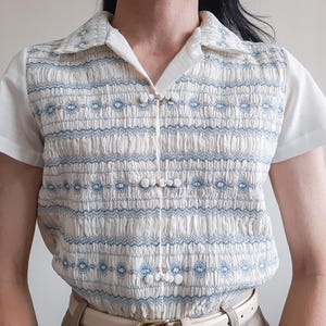 以下が含まれることがあります： 白いベースに青い刺繍模様とスモッキング加工が施された半袖シャツ。フロントには尖った襟があり、衣服の中央に縦に並んだ3つの布製ボタンで留めるデザインになっています。