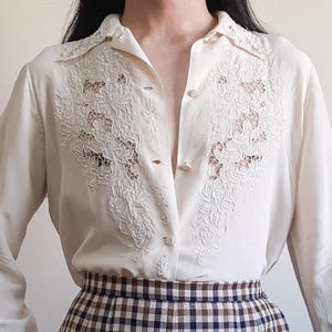 Pode incluir: Blusa creme com botões e bordado floral intrincado na frente. A camisa de manga comprida apresenta uma gola clássica e um brilho sutil. A parte inferior da imagem mostra uma saia xadrez marrom e branca.