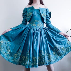 May include: Teal-colored dress and matching bolero jacket. The off-the-shoulder dress features a full skirt and is adorned with gold floral embroidery along the bodice and hem. The bolero has long sleeves and matching embroidery.