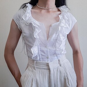 May include: A white short-sleeved blouse featuring a deep V-neckline with intricate lace-trimmed ruffles along the front placket, paired with light-colored corduroy trousers and a delicate beaded necklace worn around the neck.