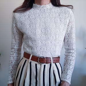 Pode incluir: Uma blusa branca de renda com mangas compridas e gola alta. É combinada com calças creme com riscas verticais e um cinto de couro castanho com fivela prateada. O conjunto é de estilo vintage.