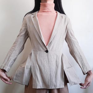 May include: A beige blazer with a subtle grid pattern, featuring long sleeves, a single dark button closure at the waist, and deep vertical side slits. The blazer is worn over a peach-colored ribbed turtleneck sweater and brown trousers.