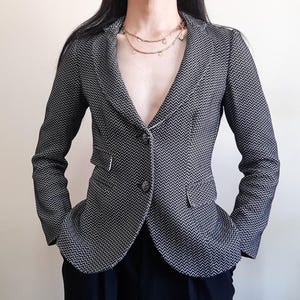 May include: A fitted blazer featuring a black and white chevron pattern, long sleeves, a notched lapel, and two front buttons. The garment includes two side flap pockets and is worn with a layered gold-tone chain necklace with small circular charms.