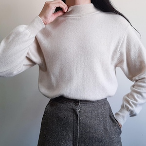 May include: A cream-coloured long-sleeve jumper featuring a mock neck collar and ribbed cuffs, paired with dark grey textured trousers. The jumper has a soft knit texture and a relaxed fit, while the trousers feature a visible front button closure.