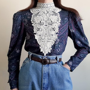 May include: A long-sleeved blouse with a paisley pattern in shades of blue, purple, and green. The blouse features a high, white lace collar and cuffs. The blouse is tucked into blue denim jeans with a brown belt.