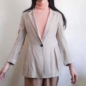 May include: A beige blazer with a subtle grid pattern, featuring long sleeves, a notched lapel, and a single dark button closure at the waist. The blazer is worn over a peach-colored ribbed turtleneck sweater with long sleeves.