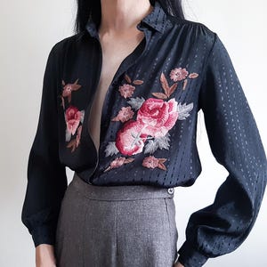 Puede incluir: Una camisa negra de manga larga con botones, un sutil patrón de rayas verticales texturizadas y bordados florales detallados en tonos rosa, rojo y gris en los paneles frontales, combinada con pantalones grises de cintura abotonada.
