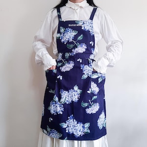 Op de afbeelding: Een marineblauwe schort met een bloemenpatroon van hortensia's in blauw en wit. De schort heeft twee zakken en de tekst "HANAE MORI" bovenaan gedrukt. Het heeft verstelbare bandjes.