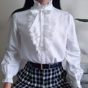 May include: A white blouse with a high ruffled collar and a delicate lace-trimmed bib. The long sleeves end in ruffled cuffs. A black belt with a gold buckle is visible. The blouse is paired with a black and white plaid skirt.
