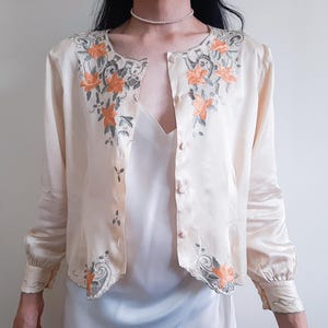 Puede incluir: Una blusa de manga larga color crema con botones, que presenta bordados florales naranjas y detalles de encaje gris a lo largo del cuello, la tapeta frontal y los puños. La prenda tiene un cuello redondo y un dobladillo festoneado.