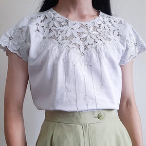 Può includere: Una camicetta bianca a maniche corte con un intricato carré in pizzo floreale che si estende sulle spalle. Il corpetto presenta delicate linee ricamate verticali con piccoli accenti floreali, abbinata a pantaloni verde chiaro con chiusura.