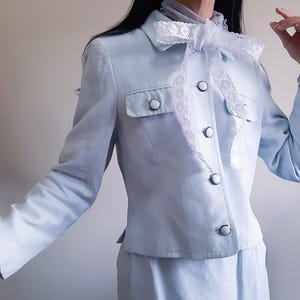 K&ouml;nnte beinhalten: Hellblauer Anzug mit passendem Rock. Die Jacke hat ein klassisches Design mit Knopfleiste, zwei Brusttaschen und einer dekorativen wei&szlig;en Spitzenschleife. Der Anzug hat eine weiche, pastellfarbene Farbe.