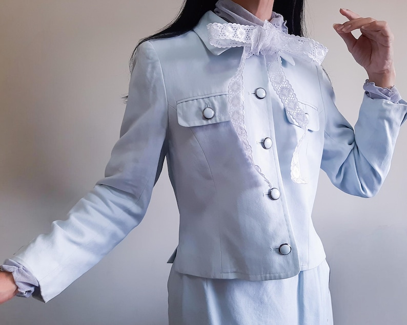 K&ouml;nnte beinhalten: Hellblauer Anzug mit passendem Rock. Die Jacke hat ein klassisches Design mit Knopfleiste, zwei Brusttaschen und einer dekorativen wei&szlig;en Spitzenschleife. Der Anzug hat eine weiche, pastellfarbene Farbe.