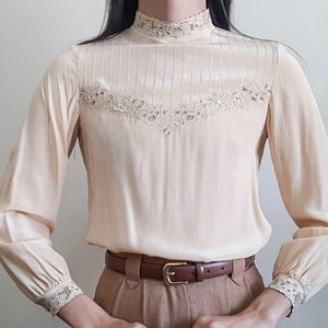 Op de afbeelding: Een blouse met lange mouwen in een bleke crèmekleur, voorzien van een hoge kraag, verticale plooien op de borst en gedetailleerde kanten afwerking langs de halslijn, manchetten en een V-vormig paneel over het bovenlijf, gecombineerd met een bruine riem.