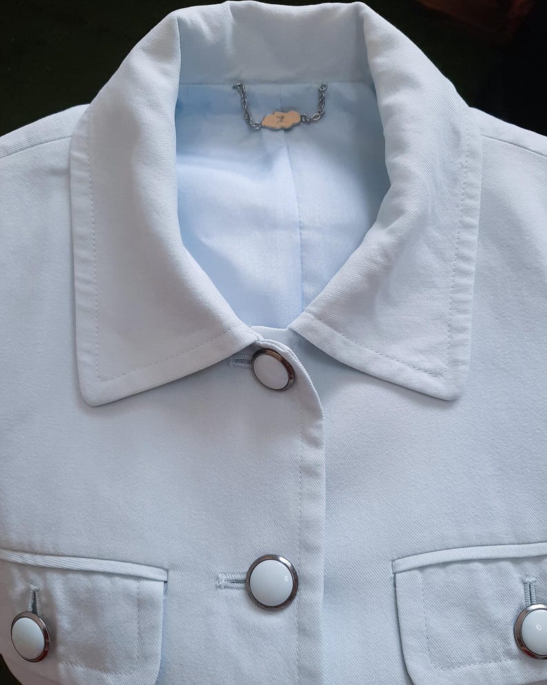 K&ouml;nnte beinhalten: Hellblaue Jacke mit klassischem Kragen, zwei Vordertaschen und wei&szlig;en Kn&ouml;pfen mit silbernen R&auml;ndern. Die Jacke hat eine taillierte Passform und ein hellblaues Futter. Eine kleine dekorative Kette ist am Kragen befestigt.
