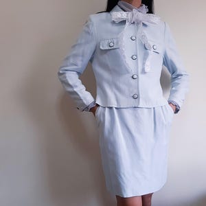 K&ouml;nnte beinhalten: Ein hellblauer Hosenanzug. Die Jacke hat zwei Vordertaschen, schwarze Kn&ouml;pfe und eine wei&szlig;e Spitzenborte. Eine wei&szlig;e Spitzenschleife ist am Hals gebunden. Der Rock ist knielang und hat Taschen.