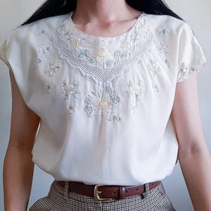 Puede incluir: Una blusa de color crema de manga corta con cuello redondo y un intrincado bordado floral en blanco y verde pálido. El diseño incluye un borde decorativo tipo encaje alrededor del cuello y delicados motivos florales en el pecho.