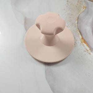 Op de afbeelding: Een lichtroze handgereedschap met een platte ronde basis en een centrale knop in de vorm van een vierbladige bloem. Het object heeft een gladde, matte afwerking en een uniforme lichtroze kleur over de gehele structuur.