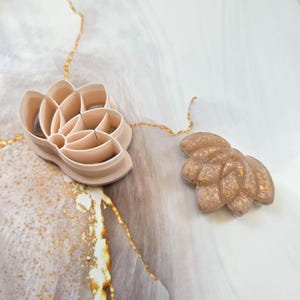Peut inclure: Un emporte-pièce en forme de fleur de lotus couleur pêche pâle avec un intérieur creux et une pièce en argile ou en pâte en forme de lotus assortie, présentant une surface texturée avec des flocons dorés incrustés, posés ensemble.