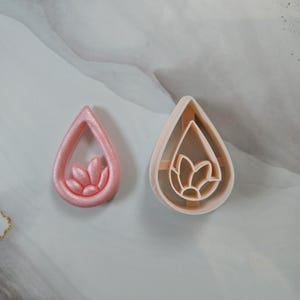 Puede incluir: Dos cortadores de galletas en forma de lágrima. Uno es de color rosa claro con un diseño de flor en el interior. El otro es de color beige claro con un diseño de flor en el interior. Los cortadores son de plástico y están sobre una superficie gris claro.