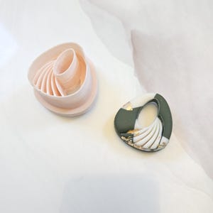 Könnte beinhalten: Ein hellrosa Kunststoff-Tonausstecher mit einem geschwungenen, gerippten Innendesign neben einem fertigen Ohrringstück mit einem dunkel olivgrünen und weißen Marmormuster, Goldblatt-Akzenten und einem ovalen Ausschnitt in der Mitte.