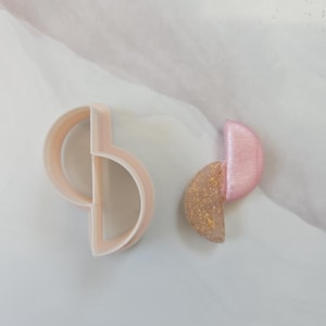 Peut inclure: Un emporte-pièce en plastique rose clair en forme de deux demi-cercles imbriqués, accompagné d'une paire de boucles d'oreilles assorties composée d'un demi-cercle rose clair uni et d'un demi-cercle translucide avec des éclats dorés.